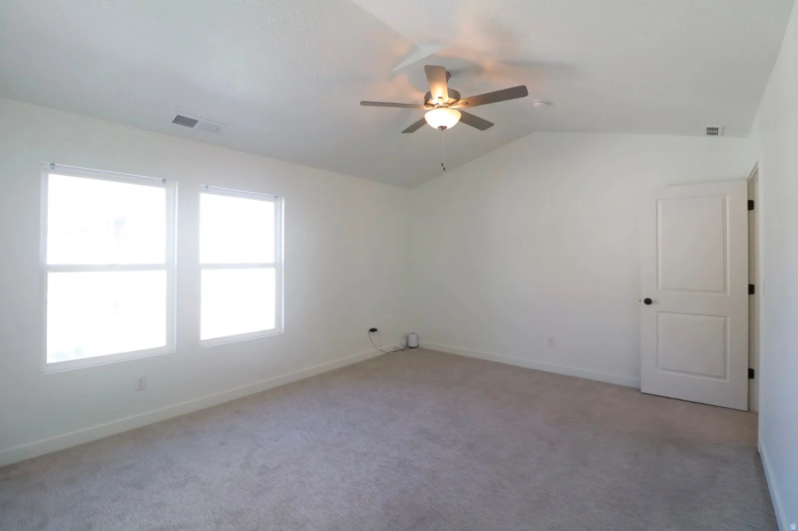 Room with vaulted ceiling, two windows, ceiling fan with integrated lighting, paneled door, and neutral-toned carpeting