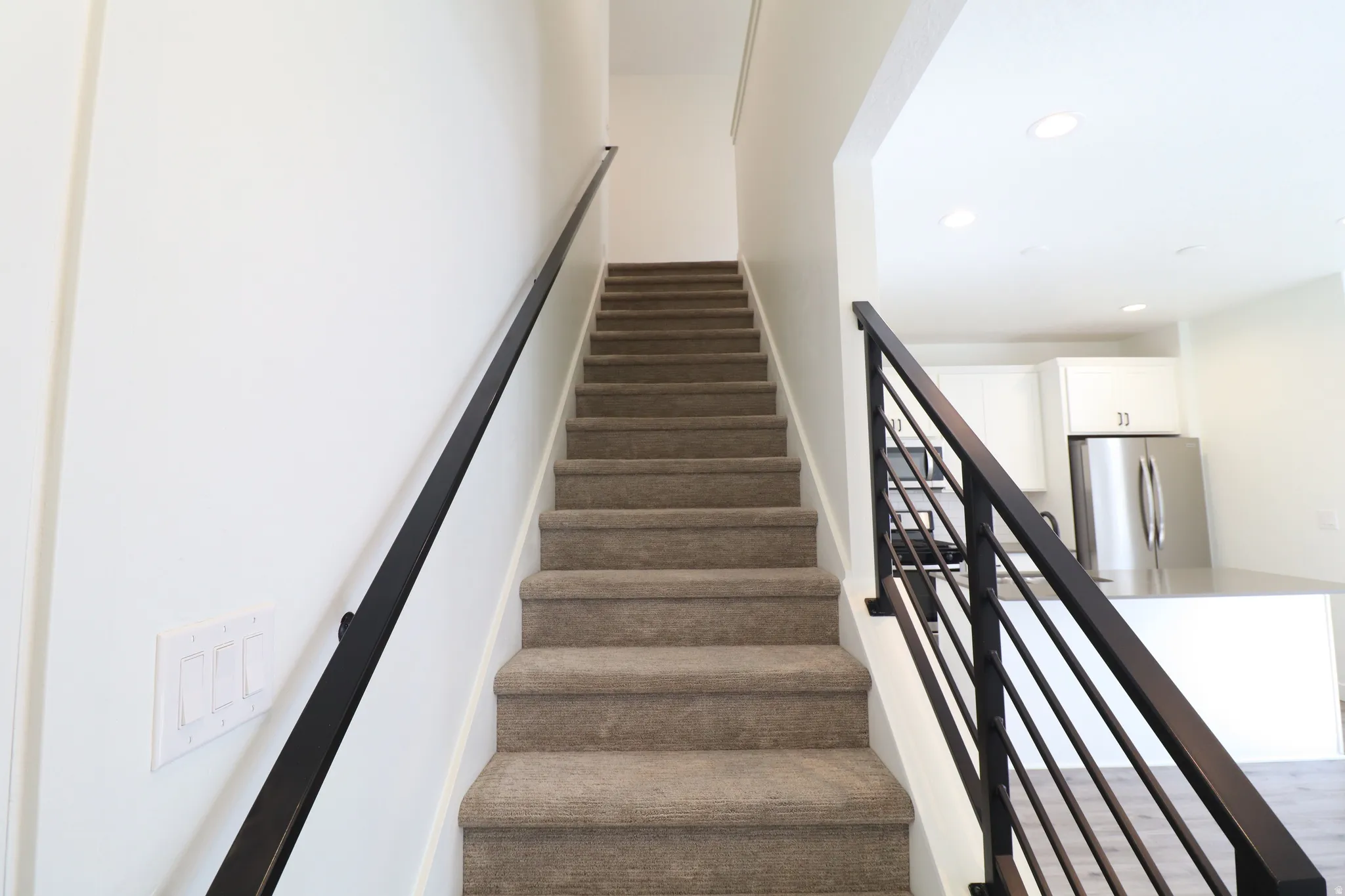 Carpeted staircase with black metal handrails and balusters, featuring a white wall and an adjacent kitchen with white cabinetry and stainless steel appliances