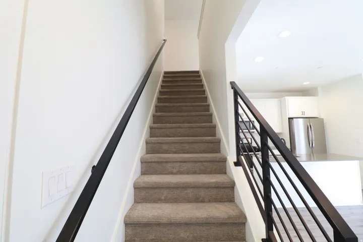 Carpeted staircase with black metal handrails and balusters, featuring a white wall and an adjacent kitchen with white cabinetry and stainless steel appliances