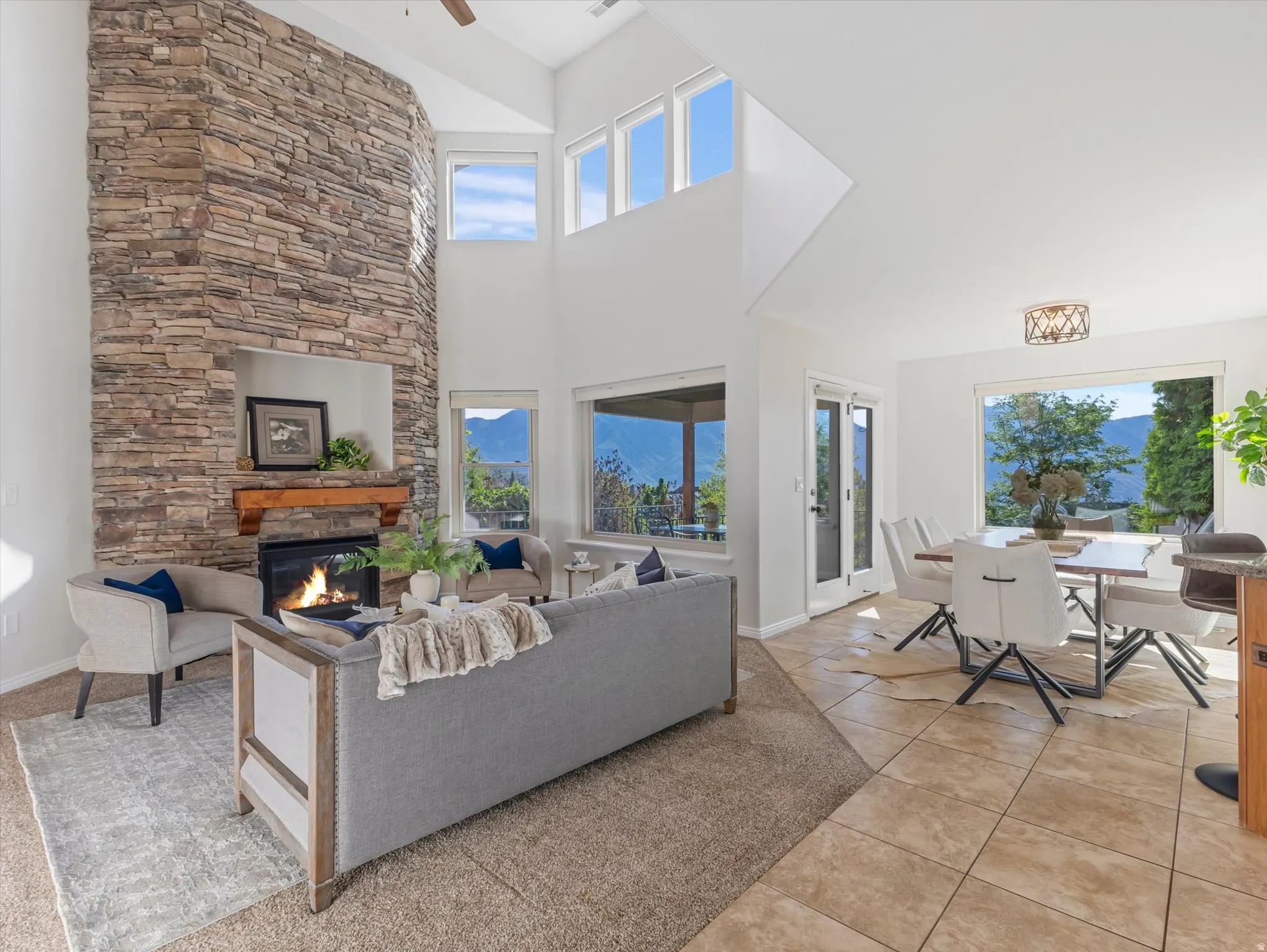 Two-story stacked stone fireplace with a wood mantle, vaulted ceilings, expansive windows, light tile flooring, and a glass-paneled door