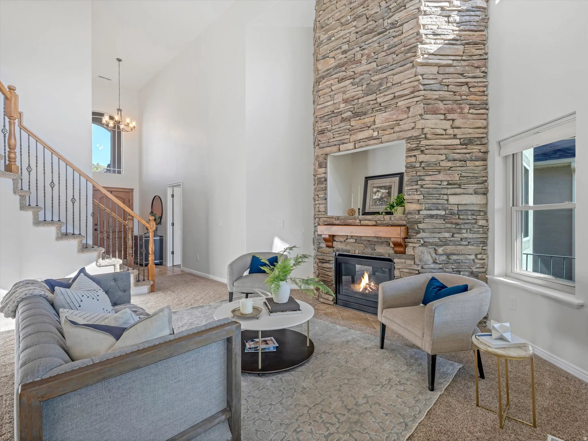 Two-story living area featuring a stone-clad fireplace with a wood mantle, a large window, a staircase with wood handrails and wrought iron balusters, and a chandelier