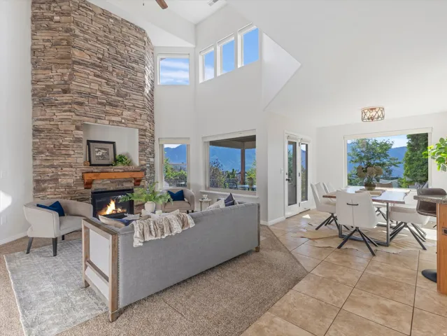 Two-story stacked stone fireplace with a wood mantle, vaulted ceilings, expansive windows, light tile flooring, and a glass-paneled door