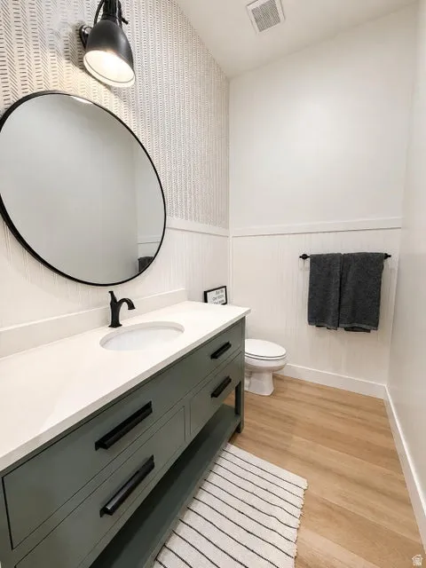 Vanity with a basin sink and matte black faucet, wall-mounted mirror, accent wallpaper, and wood-finish flooring