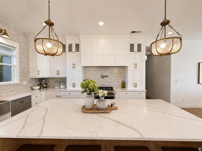 Expansive kitchen island with a light-toned countertop, white cabinetry with gold-toned hardware, full tile backsplash, and two suspended light fixtures