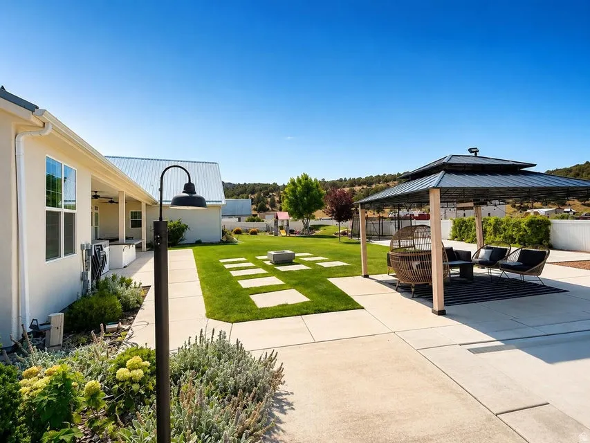 Expansive backyard featuring a shaded patio structure, concrete pathways, a manicured lawn, and planted garden beds