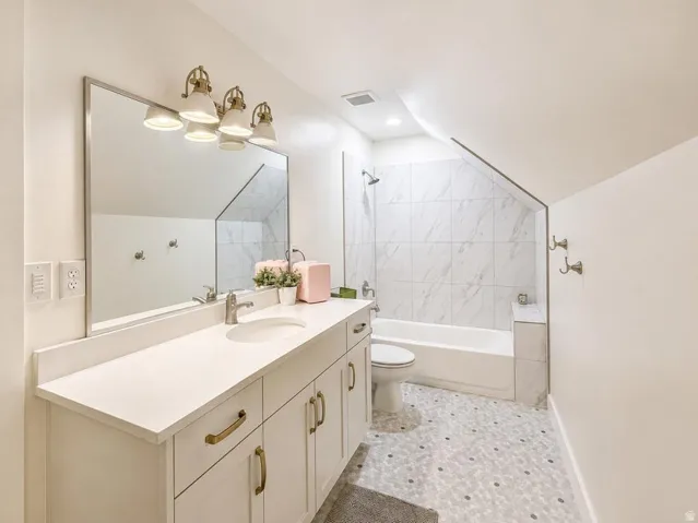 White vanity with integrated sink and brushed nickel faucet, large mirror, multi-bulb vanity light fixture, white tile shower surround with tub, and mosaic tile flooring