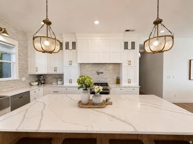 Expansive kitchen island with a light-toned countertop, white cabinetry with gold-toned hardware, full tile backsplash, and two suspended light fixtures