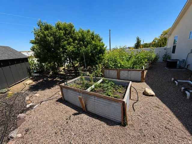 Raised garden beds with corrugated metal and wood framing, gravel ground cover, mature trees, and a detached structure with a dark roof