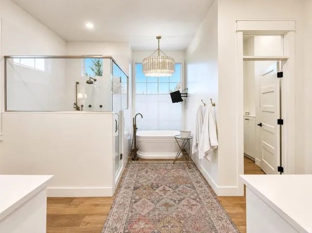 Glass-enclosed shower with multiple showerheads, freestanding bathtub, wood-finish flooring, and a decorative chandelier