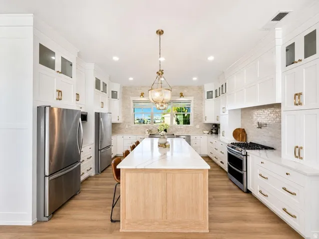 Spacious kitchen featuring an expansive center island with a light wood finish, white cabinetry with gold-toned hardware, stainless steel appliances, wood-finish flooring, and a tiled backsplash
