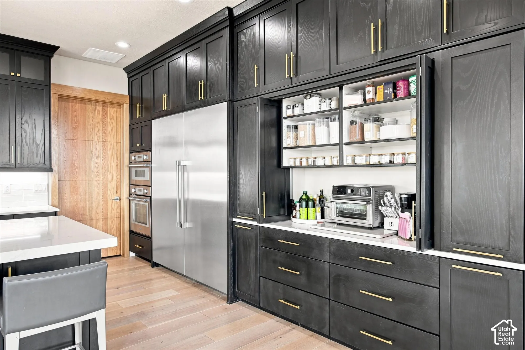 Custom black cabinetry with gold-tone hardware, integrated stainless steel refrigerator, built-in double ovens, and light wood-finish flooring