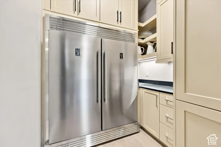 Integrated stainless steel refrigerator, cream shaker-style cabinetry, dark stone countertop, built-in shelving, wood-finish flooring