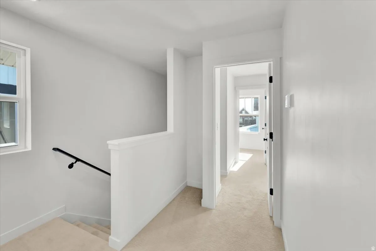 Carpeted hallway with white walls and trim, featuring a window and a staircase with a black handrail