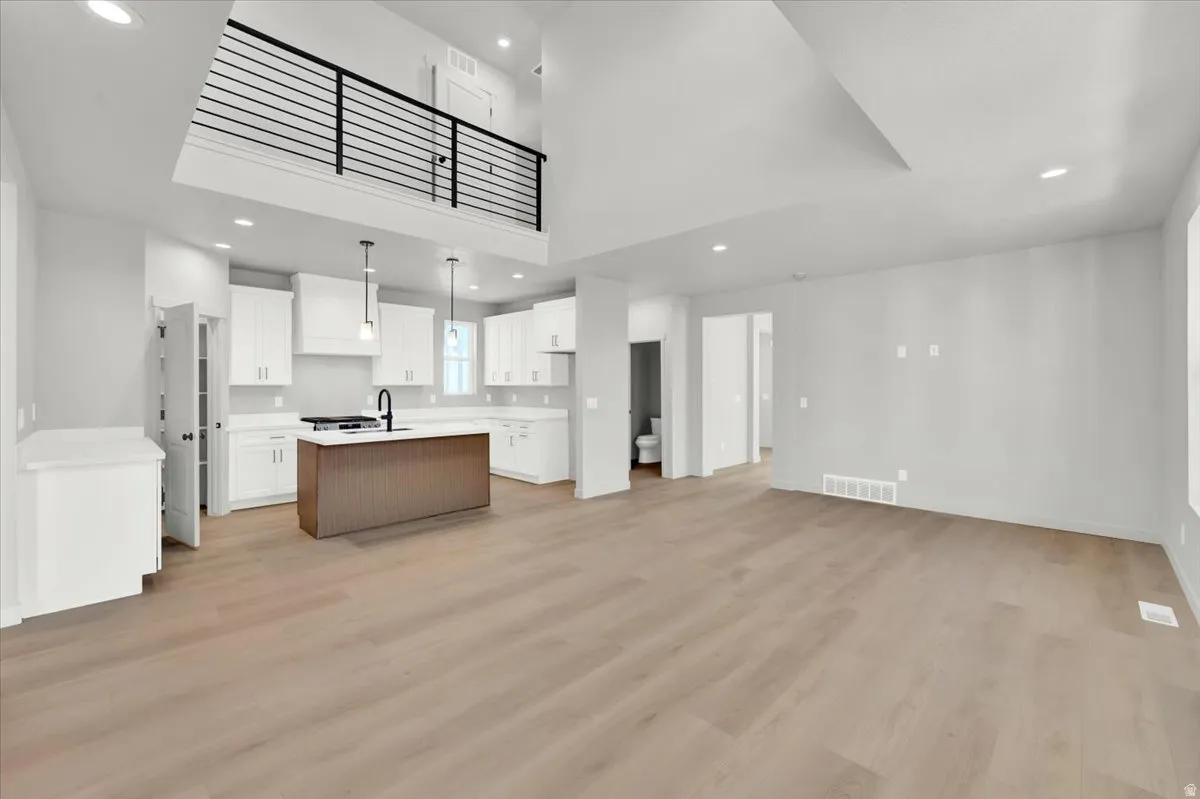 Open-concept living space featuring wood-finish flooring, a two-story ceiling, a kitchen with white cabinetry and a central island, and a second-level balcony with a metal railing