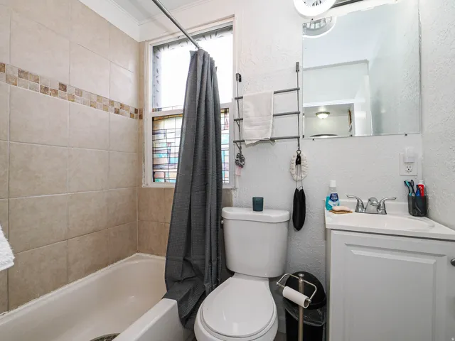Tiled shower surround with decorative accent, bathtub, window with privacy film, vanity with integrated sink, and wall-mounted mirror