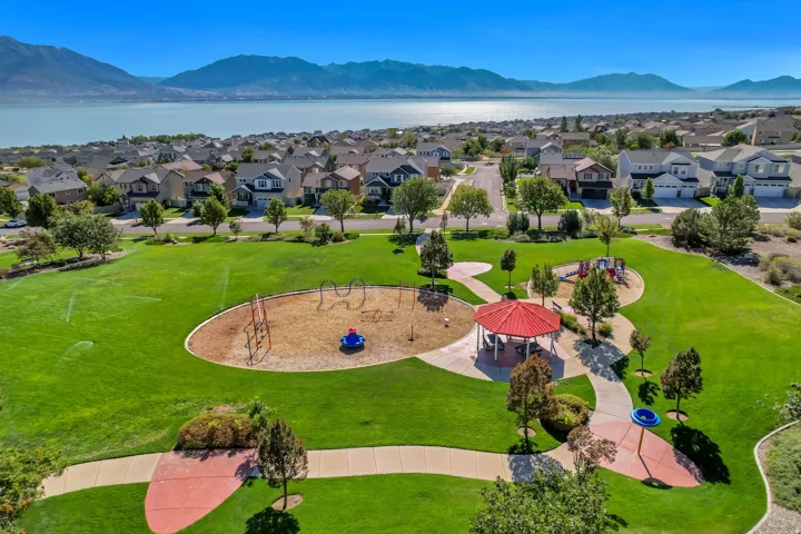 Expansive community park featuring a playground, red gazebo, paved pathways, and manicured lawns, surrounded by residential properties and a prominent view of Utah Lake with distant view of Wasatch Mountains