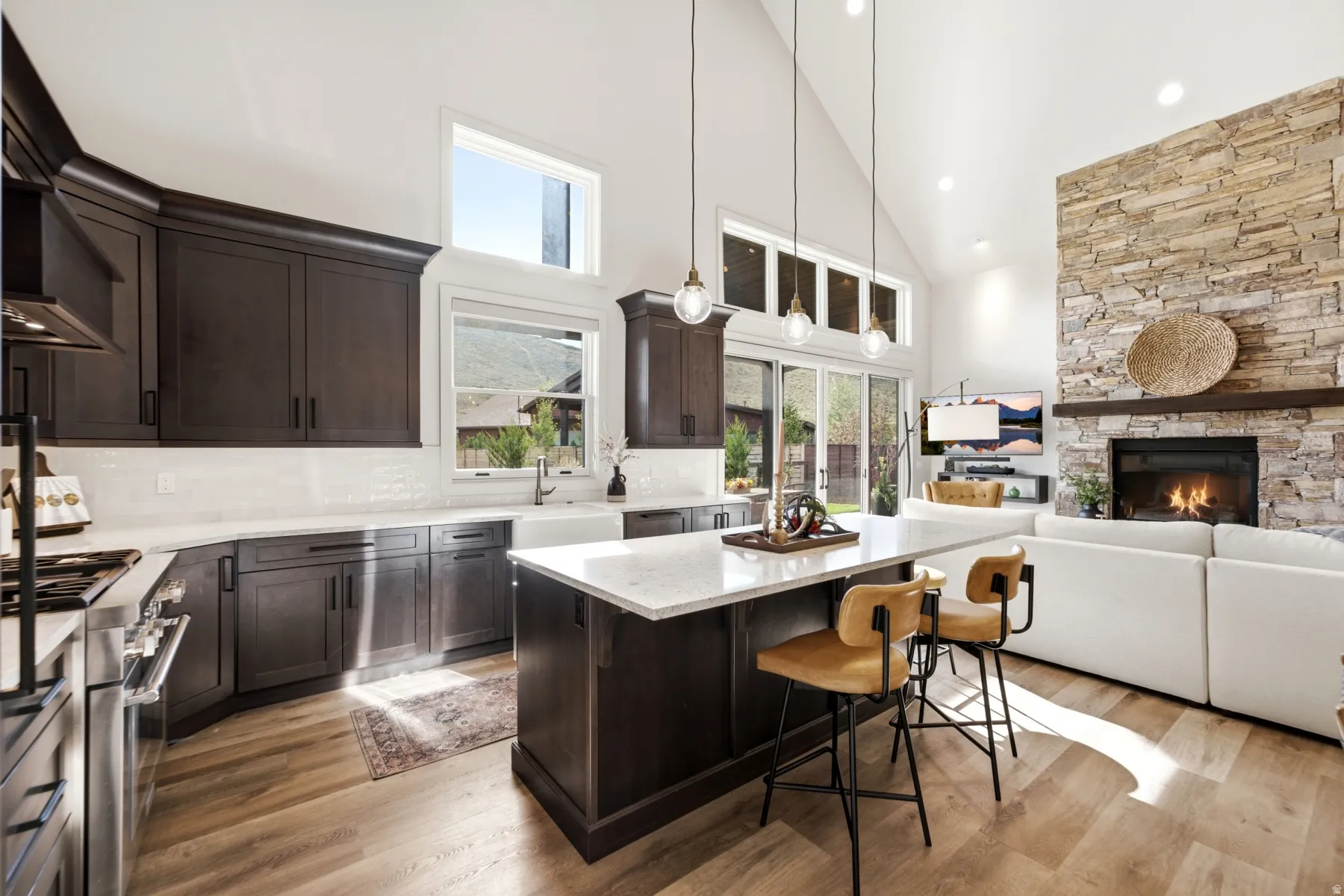 Vaulted ceiling featuring recessed lighting, extensive wood-finish flooring, a stone-clad fireplace with wood mantel, dark wood cabinetry, and a kitchen island with a light-toned countertop