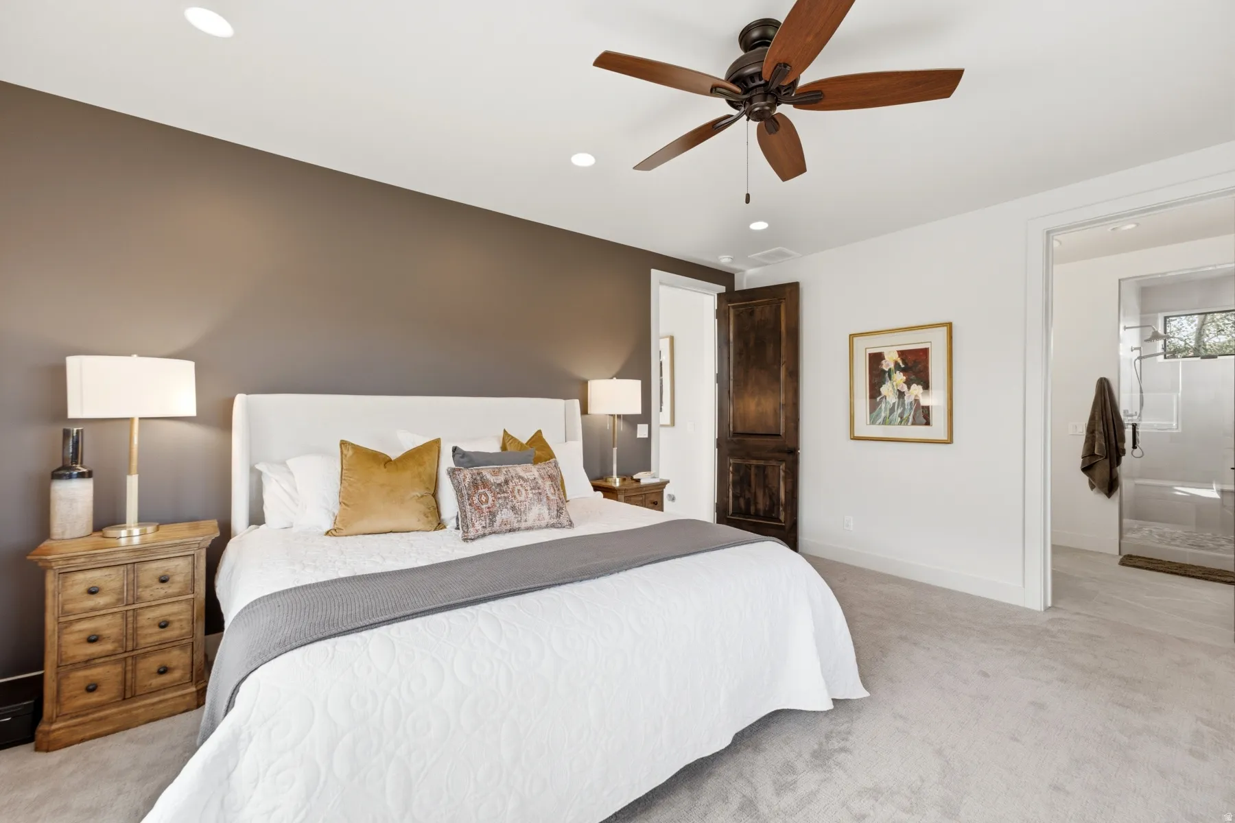 Recessed ceiling lighting, wood-finish ceiling fan, wood-finish panel door, white upholstered headboard, and light-tone carpeting