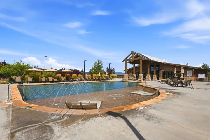 Community swimming pool with an integrated splash pad, surrounded by a paved deck and a modern clubhouse featuring a covered patio