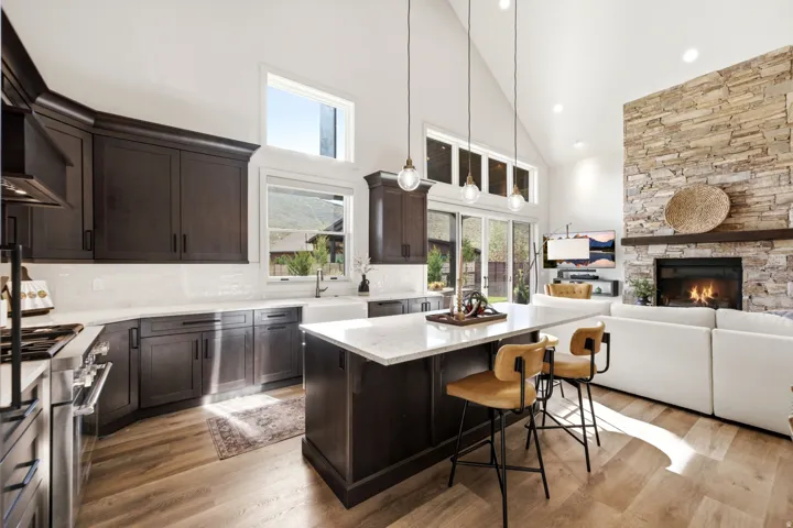 Vaulted ceiling featuring recessed lighting, extensive wood-finish flooring, a stone-clad fireplace with wood mantel, dark wood cabinetry, and a kitchen island with a light-toned countertop