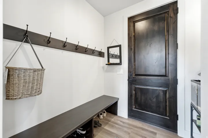 Wood-finish flooring, dark wood-finish paneled door, built-in coat rack with multiple hooks, built-in bench, wall-mounted mirror with shelf