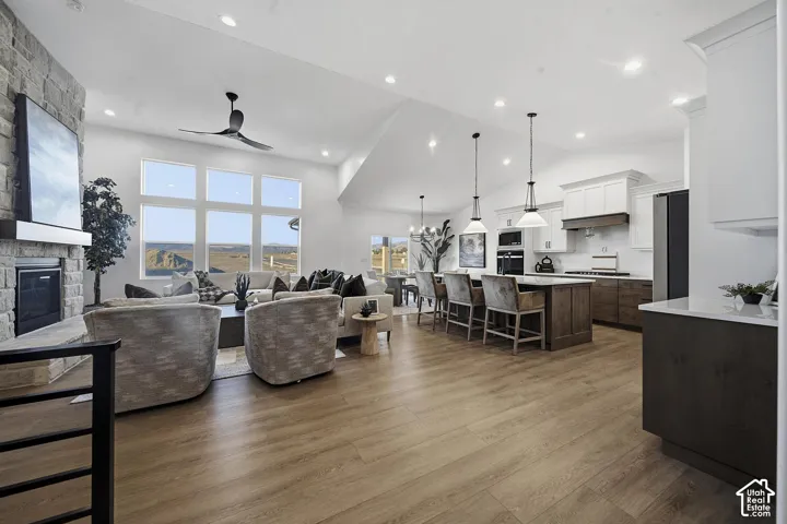 Vaulted ceiling with recessed lighting, stone-clad fireplace with mantel, expansive window wall, wood-finish flooring, and a kitchen island with pendant lighting
