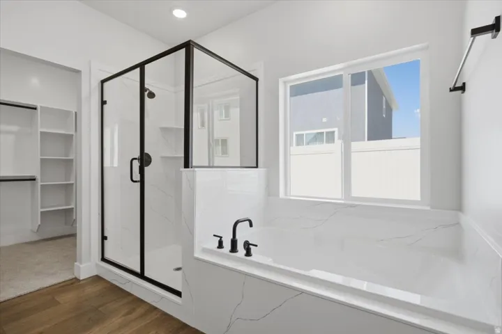 Walk-in shower with black-framed glass enclosure, built-in bathtub with black fixtures, large window, wood-finish flooring, and integrated shelving