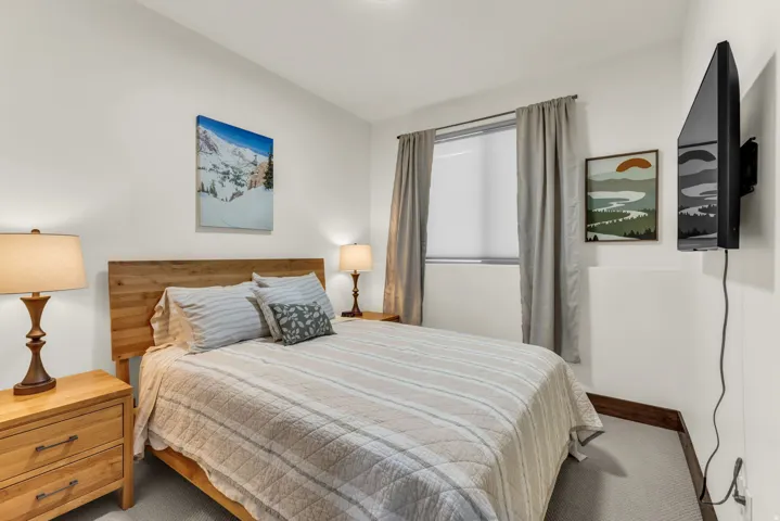 Wood-finish headboard, light gray carpet, window with blinds, baseboard trim