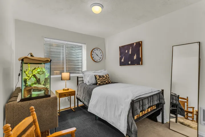 Recessed lighting fixture, light-colored walls, window with blinds, dark carpet flooring, and a metal bed frame