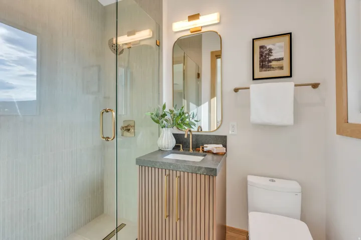 Glass-enclosed shower with gold-tone fixtures, fluted wood-finish vanity with a stone countertop, rectangular mirror with an integrated light fixture, and a white toilet