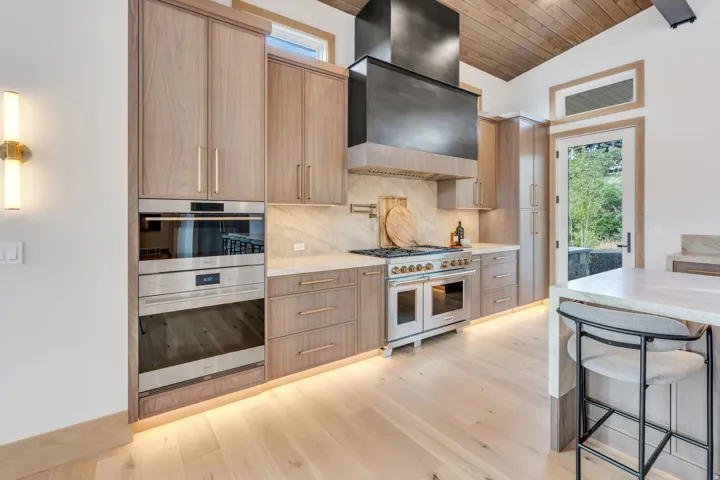 Integrated stainless steel double oven, light wood-finish flooring, wood cabinetry with gold-tone hardware, wood ceiling, and a large stainless steel range with a custom hood