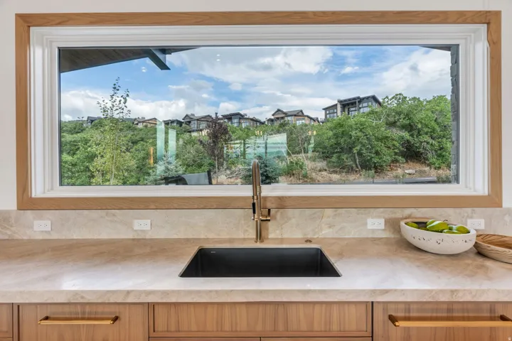 Expansive picture window with wood trim, undermount sink with brass-finish faucet, stone countertop, and wood-finish cabinetry with brass-tone hardware