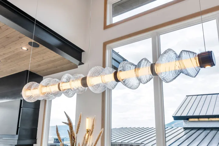 Elongated linear pendant light fixture with clear glass elements, large white-framed windows, wood-finish ceiling, and exposed black structural beam