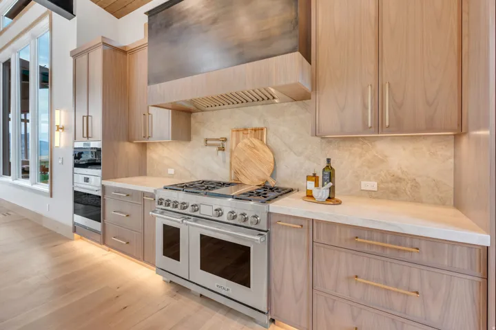 Professional-grade stainless steel range with a custom range hood, light wood-finish cabinetry, light stone countertops, and a stone backsplash
