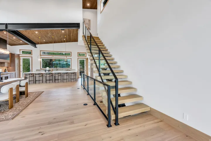 Open-concept interior featuring light wood-finish flooring, a floating staircase with glass and black metal railing, and a wood-paneled ceiling with exposed structural beams