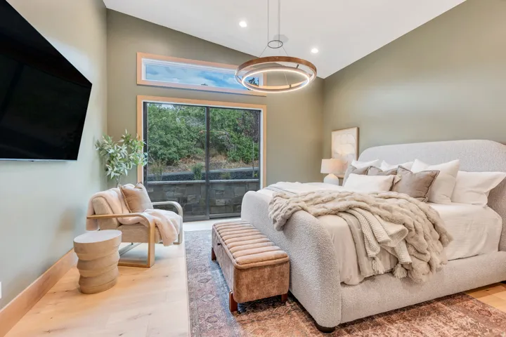 Primary Bedroom Suite with Hardwood Flooring, Sliding Glass Doors Overlooking Landscaped Grounds, Pendant Lighting, Wall-Mounted Entertainment System, and Upholstered Seating Area with Ottoman
