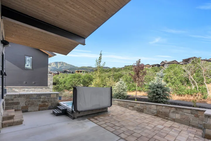 Expansive covered patio with wood-finish ceiling featuring stone veneer walls and pavers, overlooking mature landscaping and rolling hillsides
