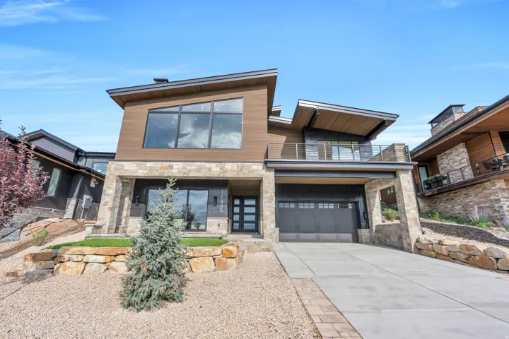 Modern architecture featuring stone and wood siding, a large two-car garage, expansive windows, and a second-story balcony with metal railings
