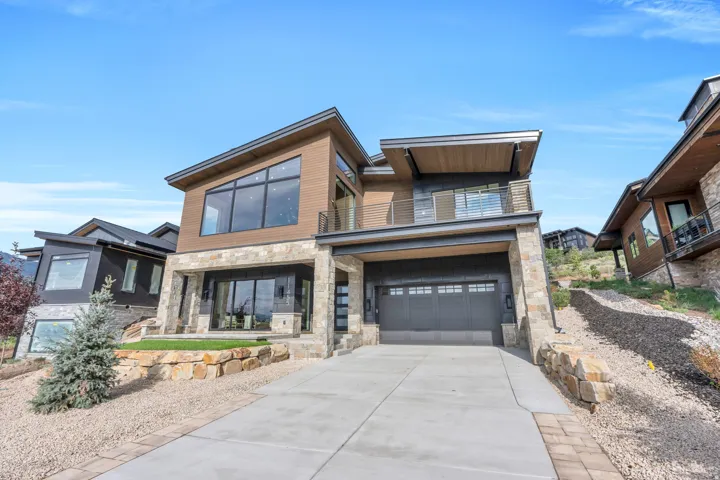 Modern exterior featuring stone and wood-finish siding, a large concrete driveway, an integrated two-car garage, and an upper-level balcony with metal railings