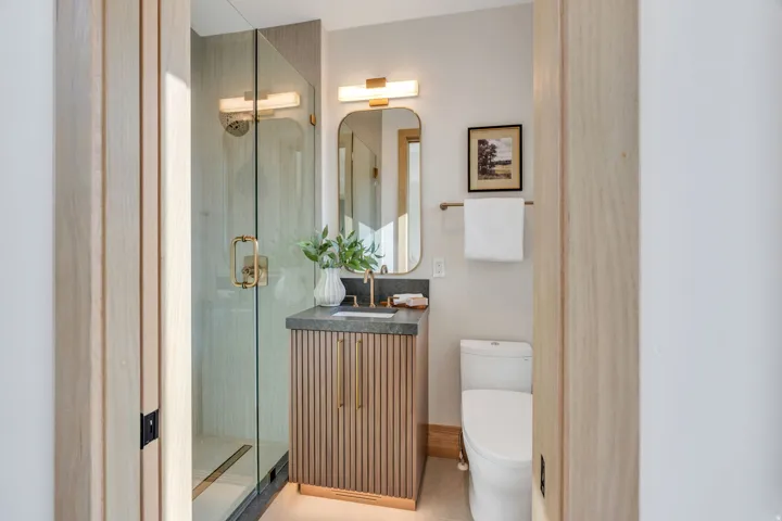 Glass-enclosed shower with gold-tone hardware, vanity with fluted wood cabinet and stone countertop, and contemporary wall-mounted mirror