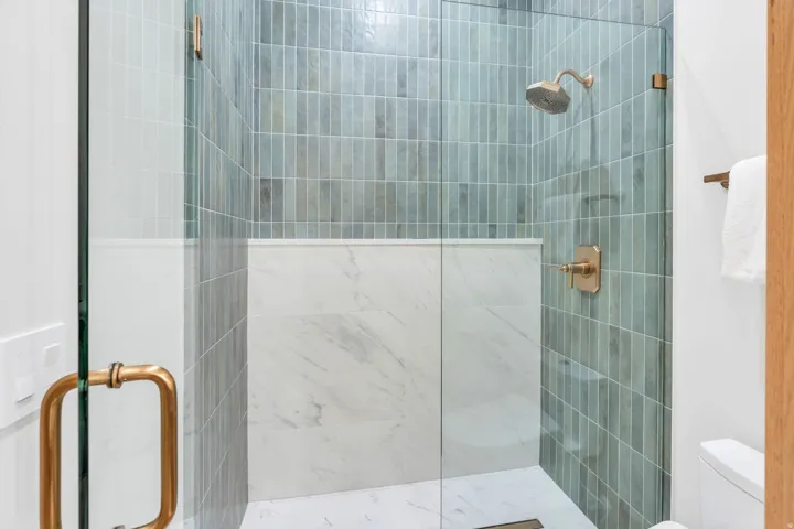 Glass-enclosed shower with vertical tile surround, marble-finish wall paneling, and coordinated bronze-finish fixtures