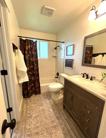 Wood-finish flooring, subway tile shower surround, dark wood vanity with white countertop, integrated sink, and matte black fixtures