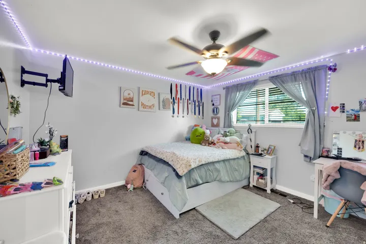 Wall-mounted television bracket, ceiling fan with integrated lighting, window with blinds and gray curtains, gray wall paint, and gray carpet flooring