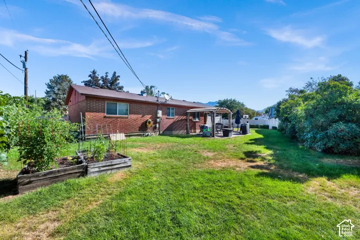 Expansive backyard featuring a brick exterior, a paved patio with a pergola, raised garden beds, and mature perimeter landscaping
