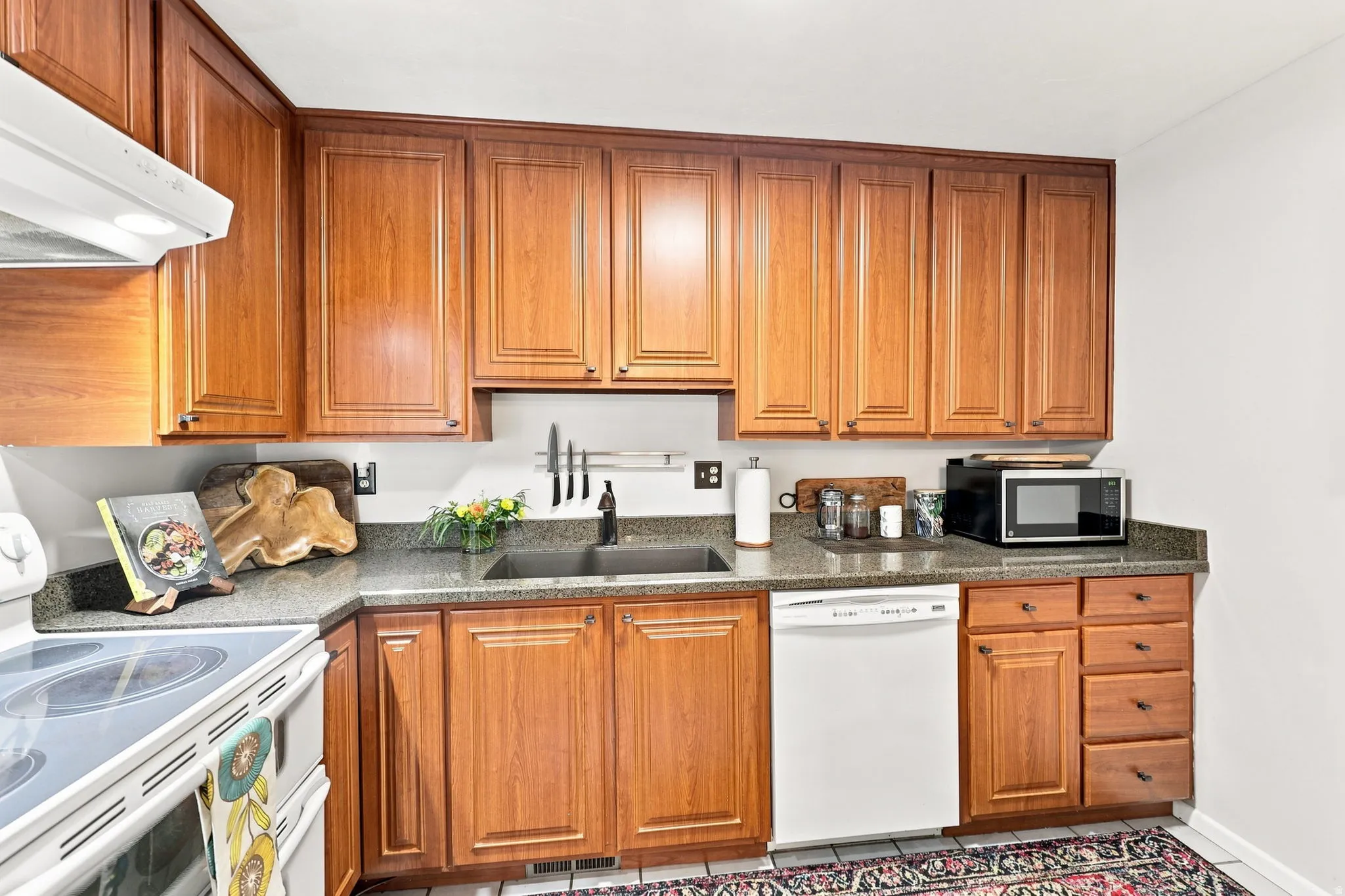 Wood cabinetry with panel detailing, stone-finish countertops, stainless steel sink, and white appliances