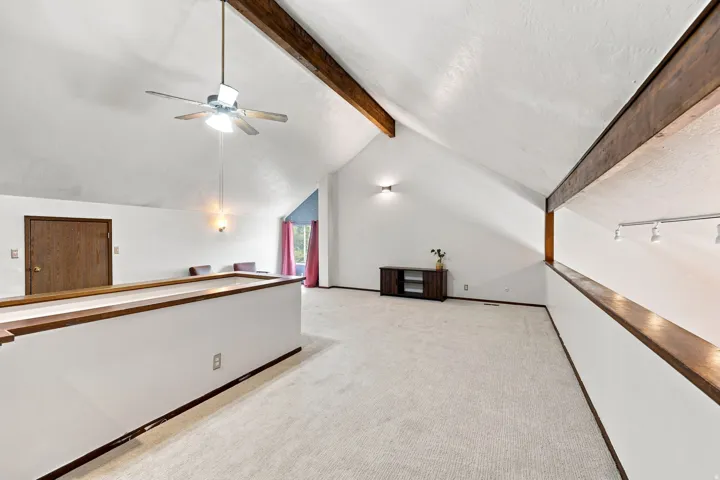 Vaulted ceiling with exposed wood beams, ceiling fan, carpeted flooring, and wood-finish railings