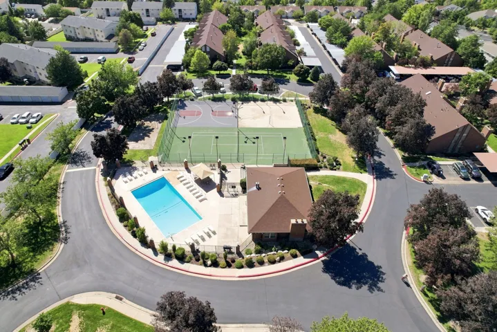 Aerial view showcasing a resort-style community amenity center featuring an illuminated swimming pool with lounge seating, multiple tennis courts with professional-grade surfacing, a sports field, mature landscaping throughout the grounds, and tree-lined