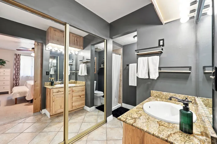 Bathroom featuring dual vanities with granite-style countertops, wood-finish cabinetry, ceramic tile flooring, and mirrored closet doors