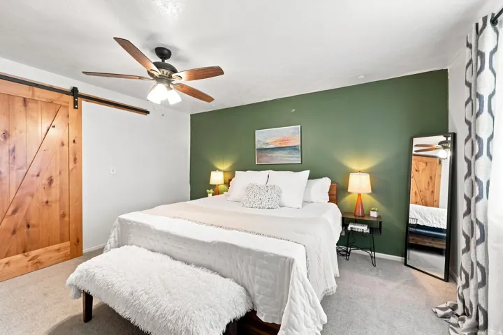 Wood barn door with black track hardware, ceiling fan with wood-finish blades, accent wall, and wall-to-wall carpeting