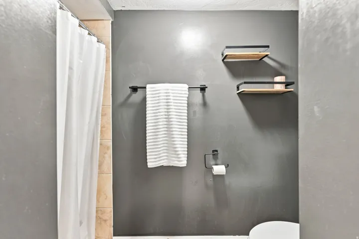Gray painted walls, black metal towel bar, black metal and wood-finish floating shelves, and tiled shower surround